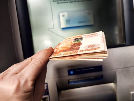 Kirov, Russia - October 08, 2024: User is withdrawing cash from an ATM, holding a stack of banknotes in a banking environment.