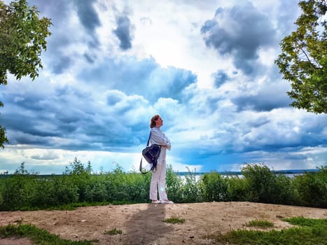Blonde woman stands thoughtfully by the river, surrounded by lush greenery and dramatic clouds on a travel adventure.
