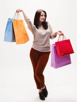 A cheerful young woman stands on white background holding shopping bags in vibrant colors. She is wearing a casual outfit and smiling, enjoying her shopping experience.