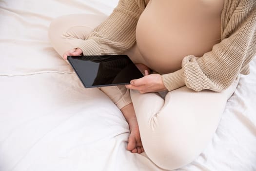 A serene and peaceful moment of a pregnant woman using her tablet while comfortably sitting on her bed