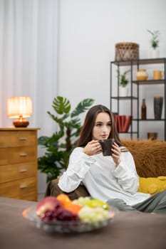 A young woman relaxes with tea in a cozy living room filled with plants and fresh fruits around her