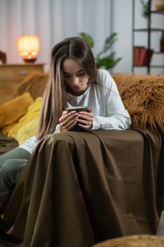 A thoughtful young woman is engaging with her smartphone while nestled in a warm, inviting living room space