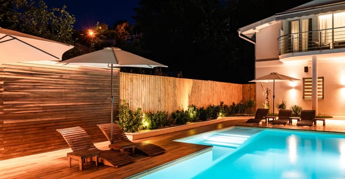 Luxury backyard with swimming pool, lounge chairs, and modern house at night. Outdoor area illuminated with soft, warm lighting