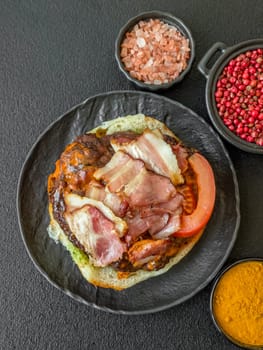 Juicy burger with beef patty, tomatoes, cheese, bacon, salt and spices on dark concrete background