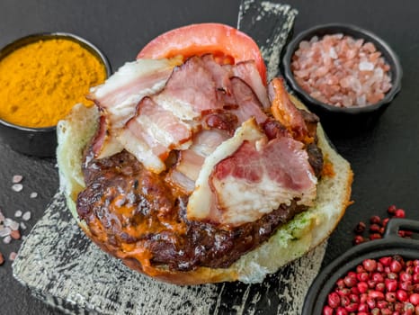 Juicy burger with beef patty, tomatoes, cheese, bacon, salt and spices on dark concrete background