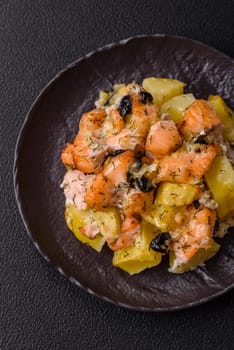 Baked salmon pieces with potatoes, cheese and olives with salt, spices and herbs on a dark concrete background