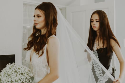 Bridesmaid adjusts the veil of a beautiful young Caucasian brunette bride holding a bouquet of white boutonnieres and looking at herself in the mirror on a summer morning, close-up side view with selective focus.
