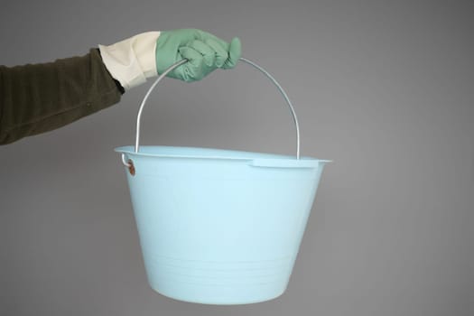 A light blue bucket is being held firmly by a gloved hand positioned against a grey backdrop