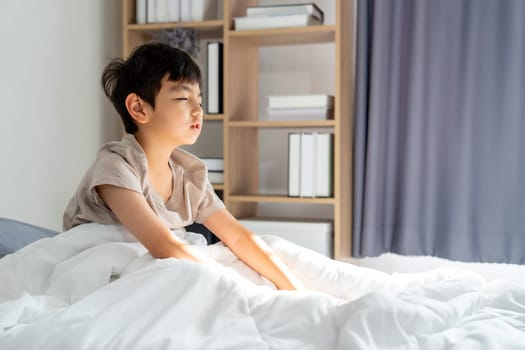 Asian boy sit on bed after wake up in the morning and his eye express asleep feeling with morning light.
