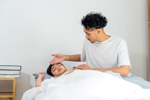 Front view Asian young father sit near his son lie on bed with sick or unwell and father take care with touch son forehead to check temperature.