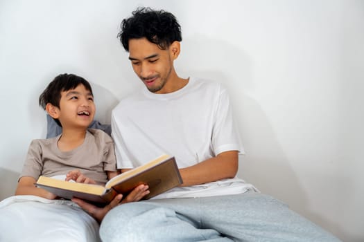 Front view Asian young father sit near his son on bed and read the book with happiness together.
