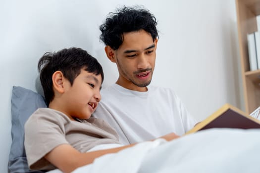 Close up view Asian young father sit near his son on bed and read the book with happiness.