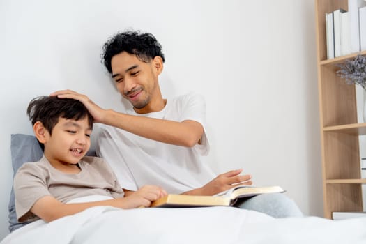 Asian young father touch head of his son during stay on bed together and read the book with happinss.