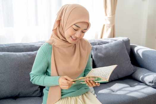 Asian beautiful muslim woman sit on sofa in her house and read the book relate to her religion and smile with happiness.