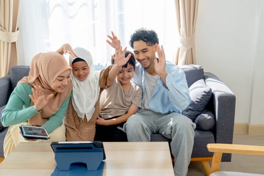 Front view of Asian muslim family compose of young mother, father, daughter and son hug and stay together on sofa in living room of their house and show greeting or say hi to tablet for online meeting