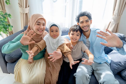 Asian muslim family compose of young mother, father, daughter and son hug and stay together on sofa in living room of their house and look at camera and show action of greeting and smile.