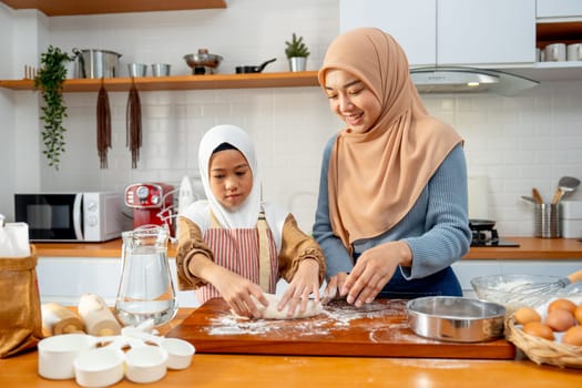 Wide shot of young Asian muslim mother help and teach her daughter to threshing flour by hands on wooden tray for making the bakery in the kitchen of their house.