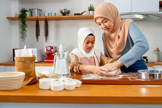Young Asian muslim mother help and teach her daughter to threshing flour by hands on wooden tray for making the bakery in the kitchen of their house.