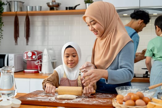 Little Asian muslim girl enjoy to use rolling pin to make some bakery and support beside by her young mother in the kitchen with father and brother also cooking in the back.