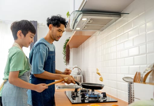Young Asian father show skill of cooking by throwing pan cake up and his son cheer up beside in the kitchen of their house.