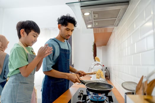 Side view young Asian father show skill of cooking by throwing pan cake up and his son cheer up beside in the kitchen of their house.