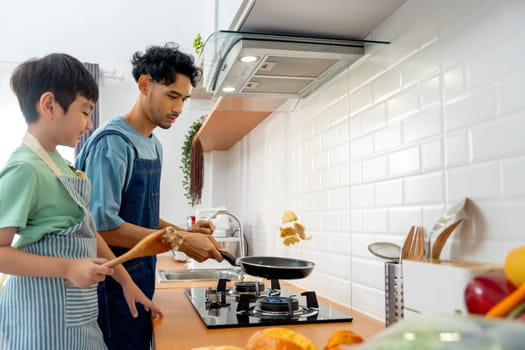 Young Asian father show skill of cooking by throwing pan cake up and his son cheer up beside in the kitchen of their house.