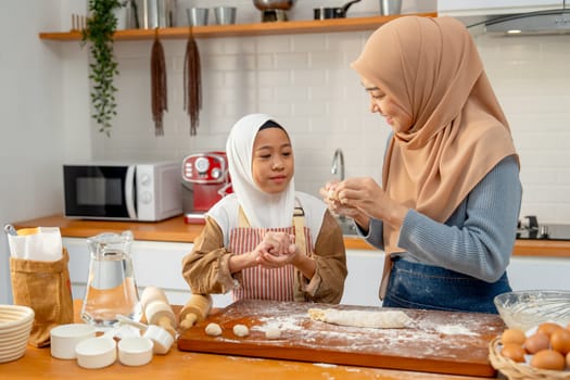 Side view of Asian muslim mother teach her daughter to threshing flour to be like ball shape for making bakery together in kitchen of their house.
