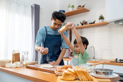 Young Asian father pour cooking flour on wooden tray during teaching his son who hold rolling pin for threshing flour for making bakery in kitchen of their house.