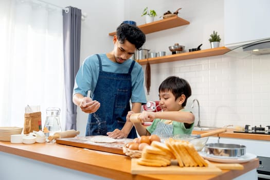 Wide shot of young Asian father pour cooking flour on wooden tray during teaching his son to thresh flour for making bakery in kitchen of their house.