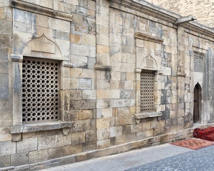 The narrow streets of Icherisheher in Baku, Azerbaijan, featuring the intricate stonework of a historic building. The warm afternoon light highlights the unique lattice windows and textured walls