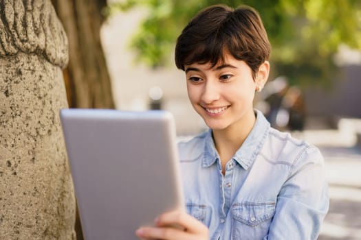A cheerful and lively young woman with short hair is smiling energetically while using her tablet outdoors, surrounded by the beautiful sights and sounds of nature all around her in the park