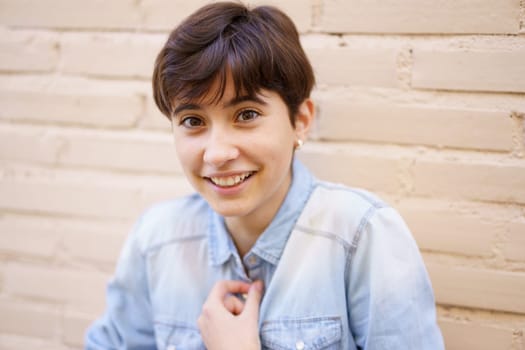 A smiling young woman with short hair, wearing a stylish denim shirt, poses playfully and joyfully against a light brick wall, radiating confidence and happiness through her vibrant expression