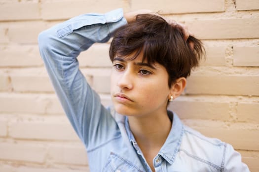 A youthful individual with a trendy short haircut, dressed in a stylish denim shirt, stands pensively in front of a roughtextured brick wall, radiating a vibe of deep contemplation and unique style