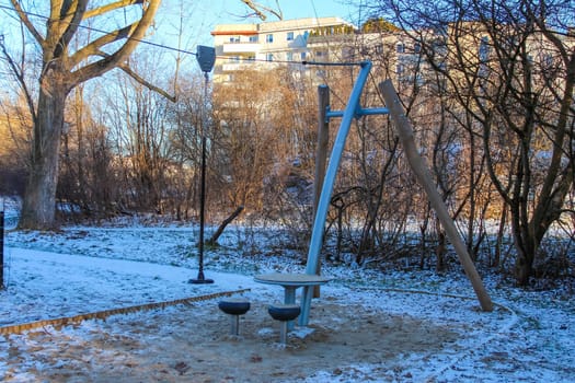 A swing set and a circular seating area stand in a snowy park surrounded by trees and sunlight in winter.