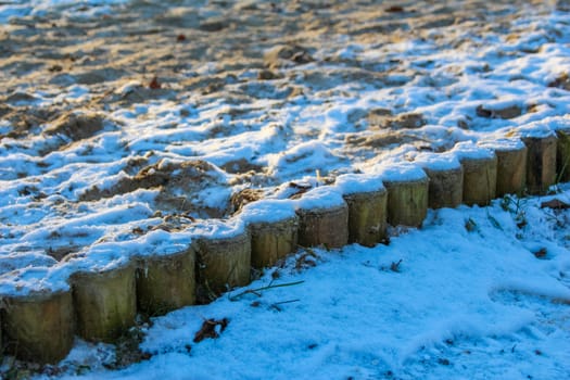 A layer of snow blankets sandy soil along a wooden border during a sunny winter day, creating a serene outdoor setting.