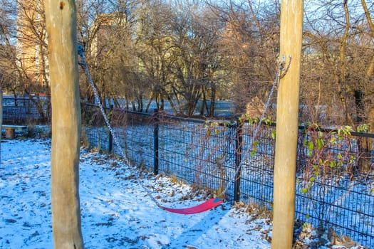 A swingset sits in a snowy park surrounded by leafless trees and a river, evoking a serene winter atmosphere.