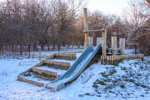 A wooden slide sits on a snowy playground surrounded by trees, inviting children to play on a crisp winter day.