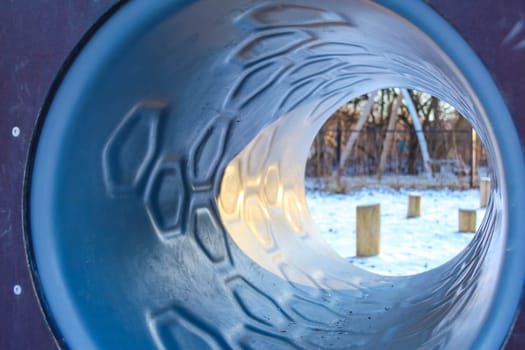 A playground tunnel gives a unique view of the snowy landscape, framed by wooden posts that enhance the tranquil winter vibe.