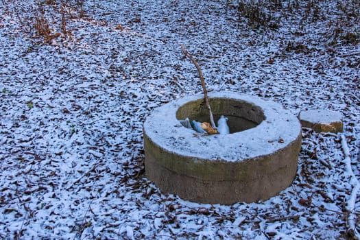 A stone well is partly covered in snow, with scattered autumn leaves nearby and a solitary stick resting inside, set in a tranquil woodland.