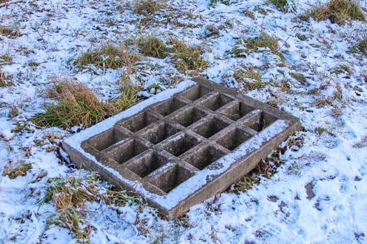 A weathered concrete slab with a grid pattern is surrounded by patches of snow and grass, revealing signs of abandonment in winter.