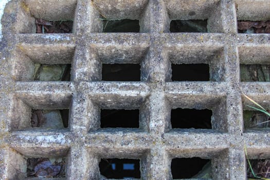 A close-up of a concrete drainage grate reveals a detailed grid pattern and a glimpse into the underground. Nature surrounds this structure.