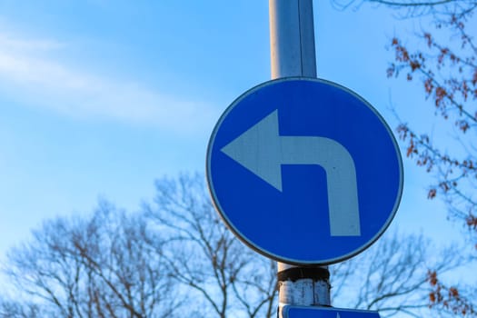 A clear blue road sign indicates a left turn in an urban area, surrounded by bare trees under a bright sky.