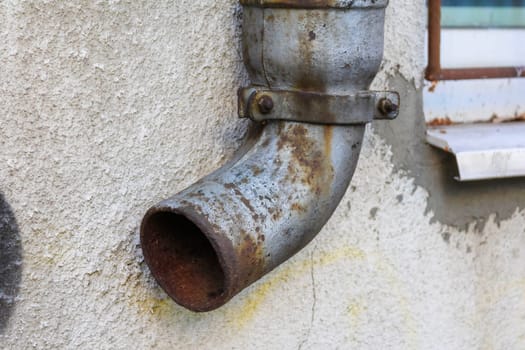 A worn metal pipe curves from a weathered wall, showing signs of rust and neglect in an urban environment.