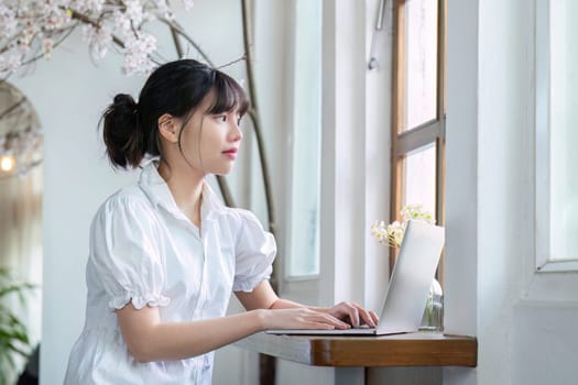 Asian female student studying online in living room with laptop doing online research for a class subject Make notes for essay homework. online education concept.