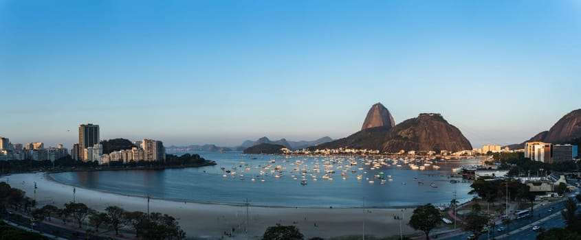 Beautiful dusk view of Botafogo, Rio de Janeiro, with highlights like Sugarloaf Mountain, Urca Hill, sailboat-filled bay and an empty beach. Ideal for text placement.