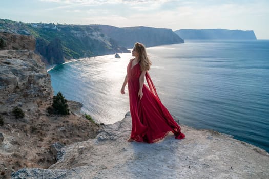 A girl with flowing hair in a long red dress stands on a rock above the sea. The stone can be seen in the sea. Sunny path to the sea from the sun