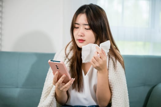Asian young woman sick sitting down on sofa with cold blowing her nose and checking smart mobile phone to cell to doctor online, depressed sad sick female holding tissues and smartphone in living room