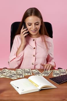 pretty woman counts money and talking by phone while sitting on chair at desk on pink background