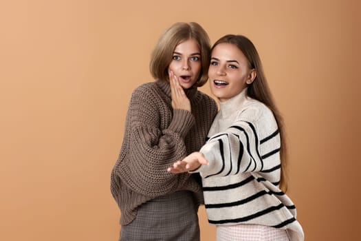 Two women friends together in casual clothes point index finger aside on beige background. Surprised blonde woman and her friend