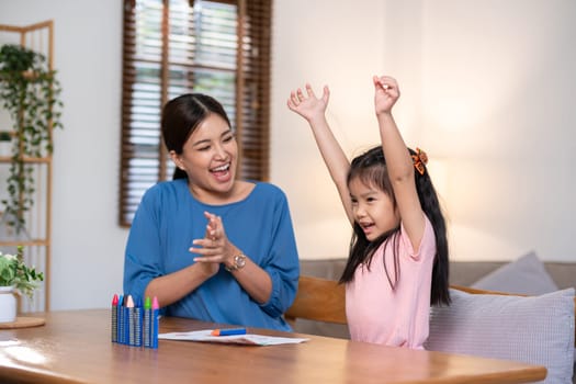 A young family spends their free time together in the living room at home. Mother and little daughter draw pictures with crayons on paper, smiling happy..